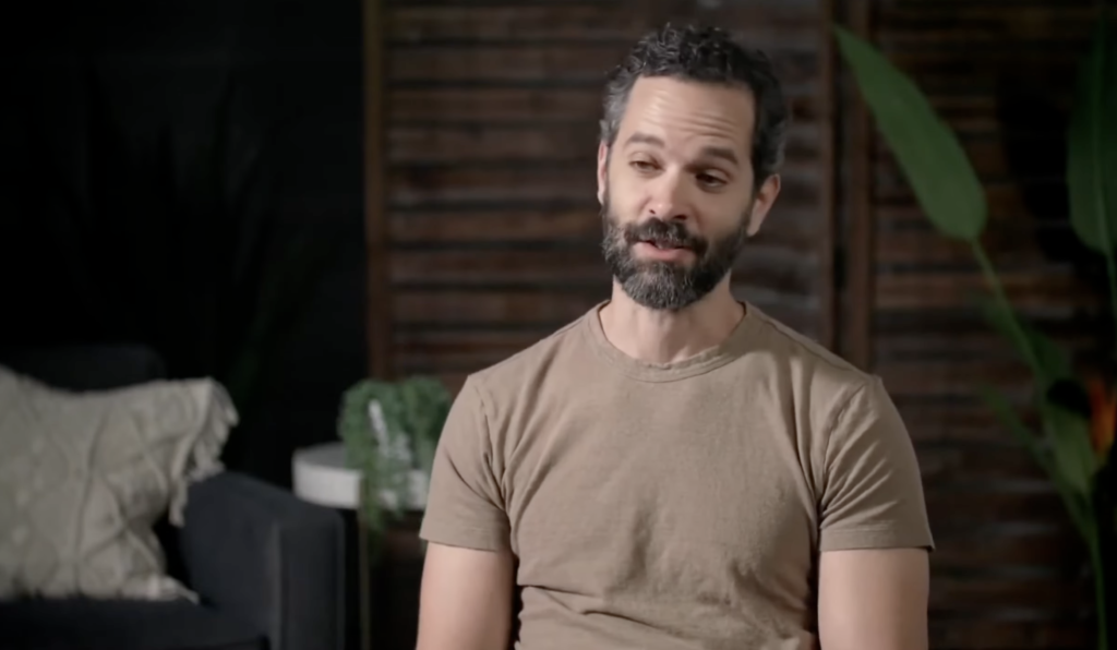 Bearded man speaking indoors near couch and plant